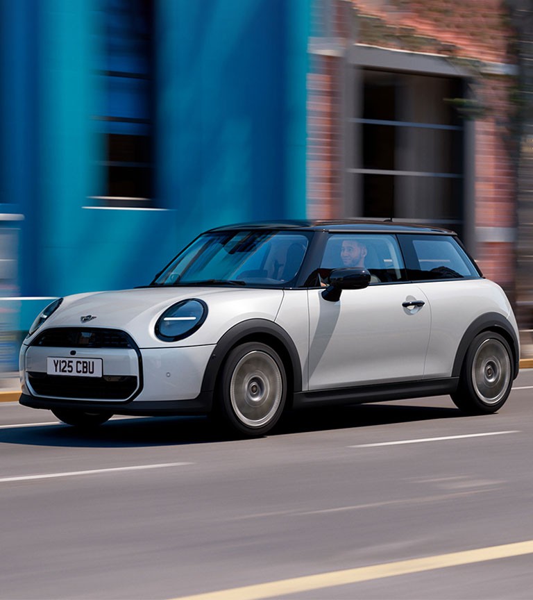 A sleek white MINI Cooper speeds along a sunny street, with colourful buildings in the background, showcasing urban life.